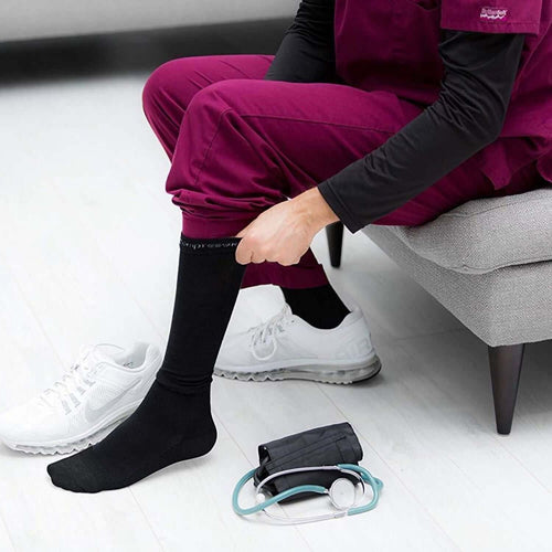 Healthcare professional wearing black compression socks (30-40 mmHg) while sitting on a couch next to a stethoscope and blood pressure cuff.